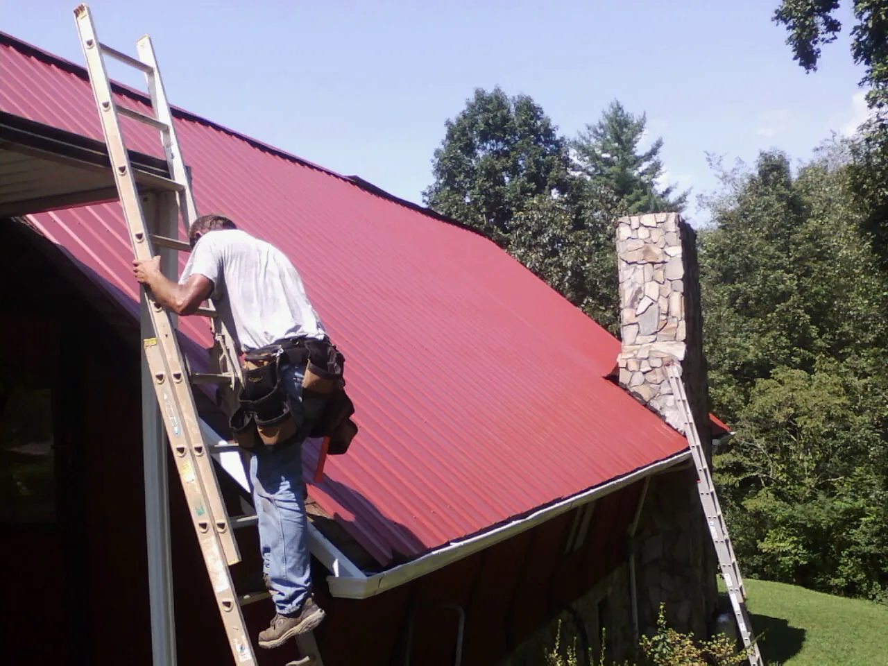 Roofer on a ladder beside a red metal roof and stone chimney — Soffit & Fascia Repair by Roofing Brothers in Stillman Valley